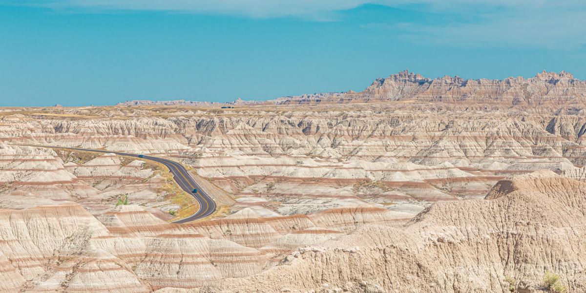 Scenic view of layered rock formations of Badlands National Park under a blue sky. A winding road cuts through the terrain, evoking isolation and adventure.