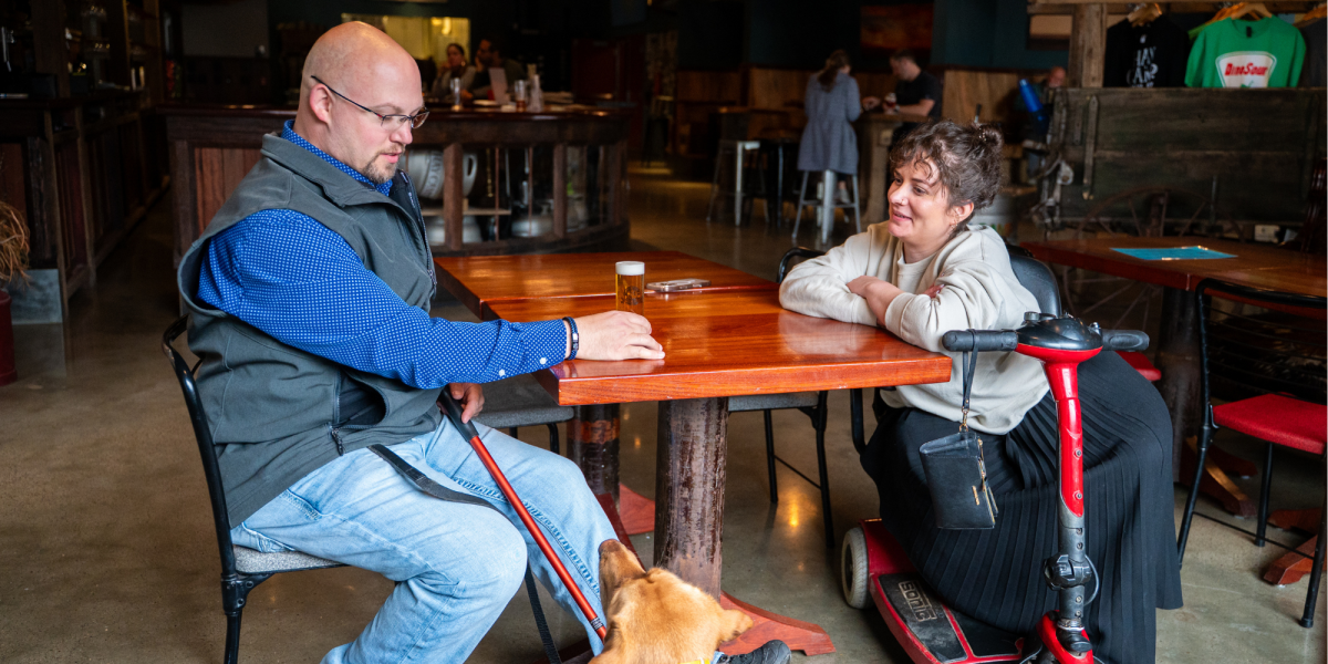 A man with a guide dog and a woman on a mobility scooter sit at a wooden table at Hay Camp Brewery, engaged in friendly conversation, evoking warmth.