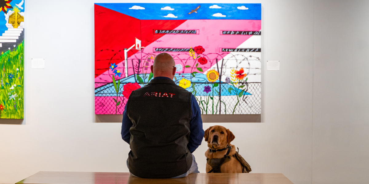 A person and a service dog sit in front of a vibrant painting. The artwork features vivid colors, flowers and barbed wire, creating a striking contrast.