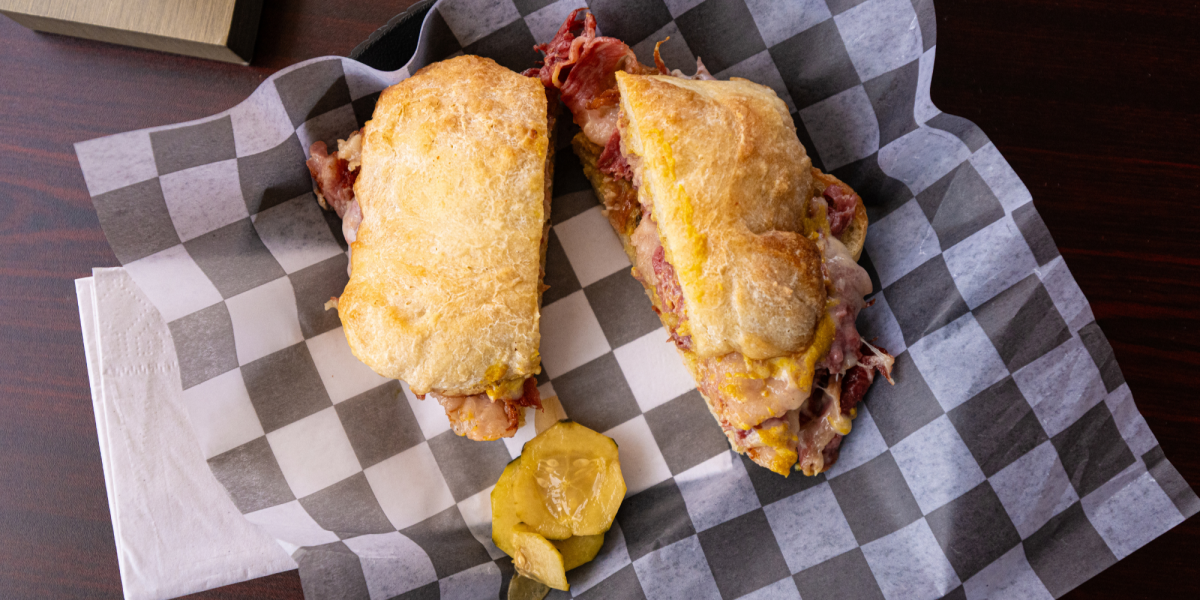 A sandwich with corned beef or pastrami on checkered paper, accompanied by pickle slices. The bread is golden brown, evoking a warm, appetizing tone.