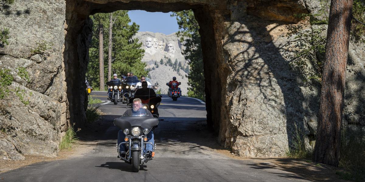 Group of motorcyclists drive through a rock tunnel with Mount Rushmore visible in the background. The scene is lively, with sunny skies and lush trees.