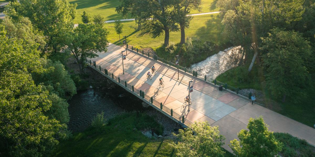 Aerial view of cyclists on a bridge over a stream, surrounded by lush trees and green fields. Warm sunlight casts long shadows, creating a serene atmosphere.