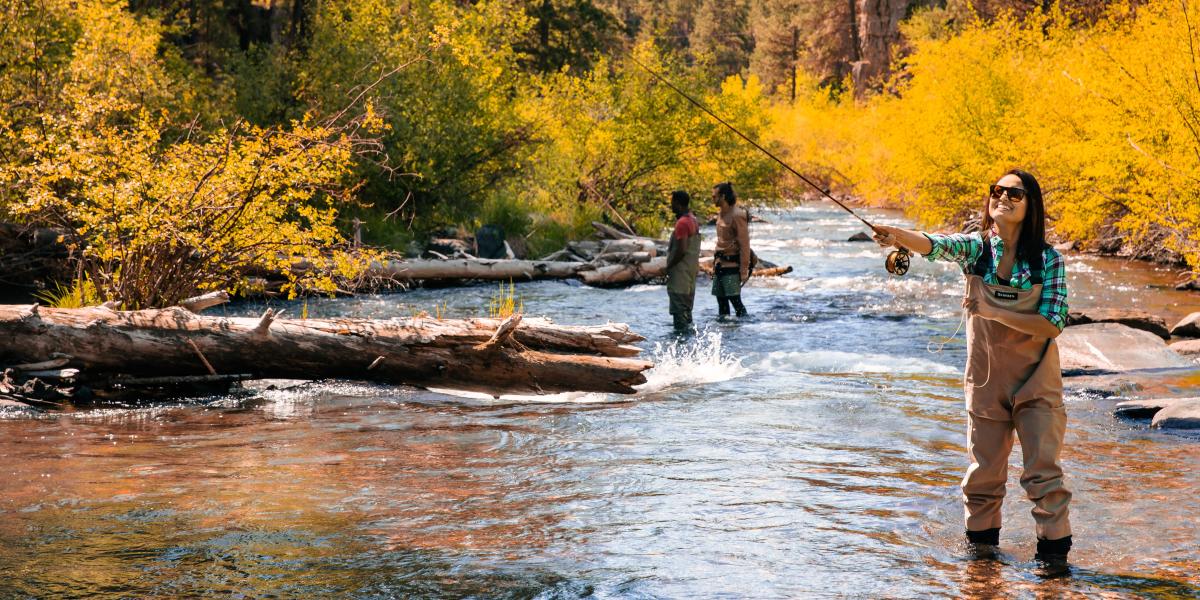 A woman in waders fly-fishes in a sunlit river, surrounded by vibrant yellow foliage. Two people stand in the background; the scene conveys tranquility and leisure.