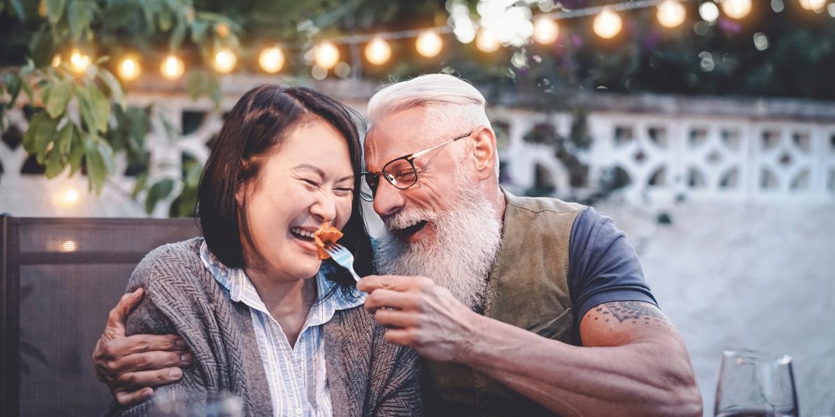 Man and woman laughing at outside dining table as he tries to feed her pasta on a fork