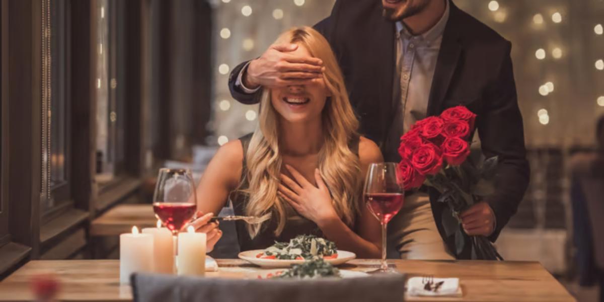 young man covering woman's eyes holding red roses at dining table
