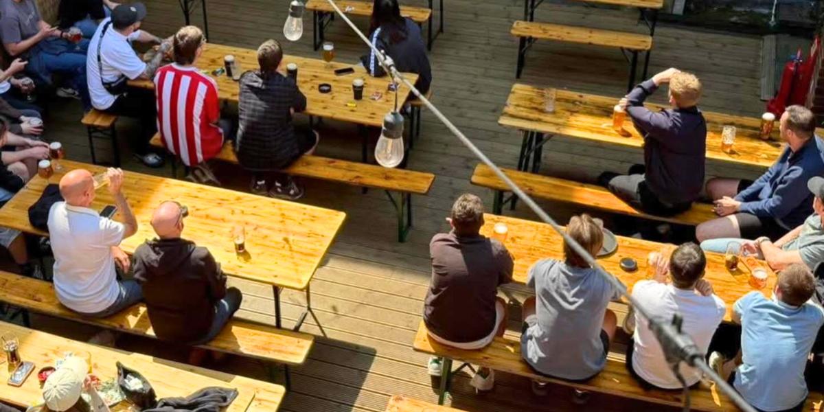 Groups of men sat on picnic benches watching football on a tv screen at The London Road Brewhouse rooftop bar