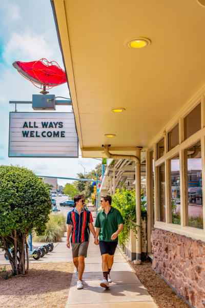 Two men walk down the sidewalk in front of Joann's Fine Foods, holding hands under a marquee sign reading "All Ways Welcome" with a neon pair of lips at the top.