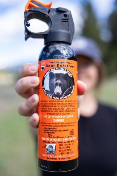 A person holds a canister of bear deterrent spray, featuring a black bear image and safety instructions on an orange label. The background shows a natural outdoor setting.