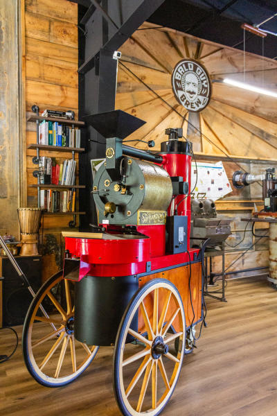 A bright red San Franciscan roasting machine sits on the warm wooden floors of a café. A black and white circular logo reading "Bald Guy Brew" can be seen hanging on the wall behind the machine.