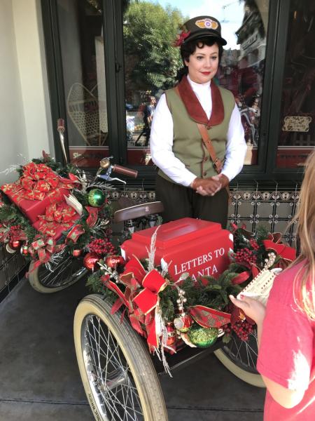 A woman is dressed in 1920s clothing and standing next to an old-timey bike that has a box in the front seat that reads 'Letters to Santa."