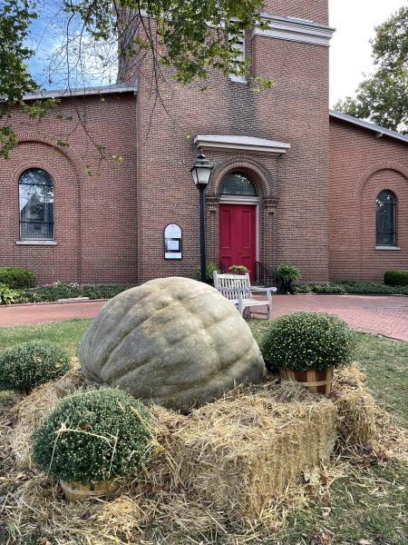 A Giant Green pumpkin in top of a hay bale in front of a church.