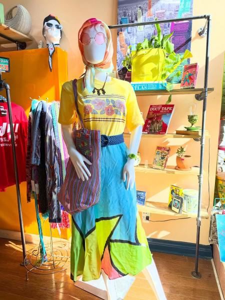A colorful mannequin display inside Mood in downtown Jeffersonville features a yellow floral graphic T-shirt, a bright patchwork skirt, a patterned shoulder bag, a headscarf, and pink sunglasses.