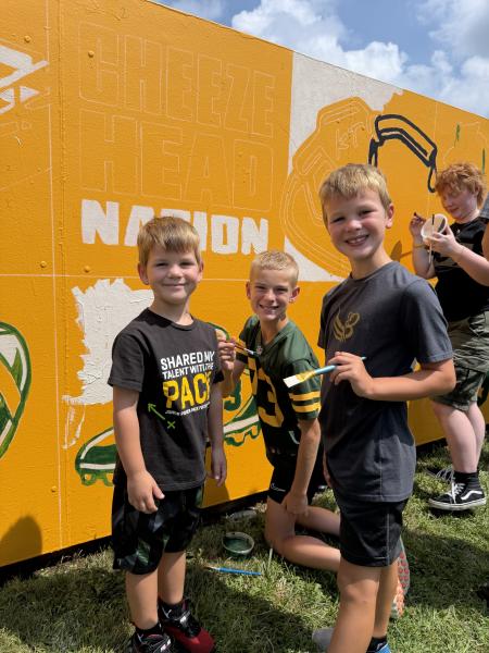 Three young boys smiling in front of the Green Bay community fence they are painting. The fence is a yellow paint by number and they are painting a tennis shoe.