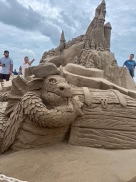 Sand sculpture with a stout dragon holding on to a treasure chest in the foreground and a traditional sandcastle in the background.