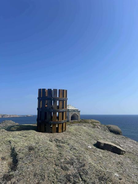 Markøy fyr med ny skulptur av kullbluss i metall plassert i forgrunnen. Fyret og skulpturen står på en bergknaus med utsikt over havet under blå himmel.