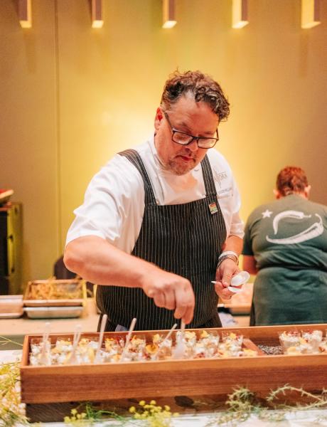 A chef in a white overcoat and pinstripe apron prepares dozens of small bites for a tasting.