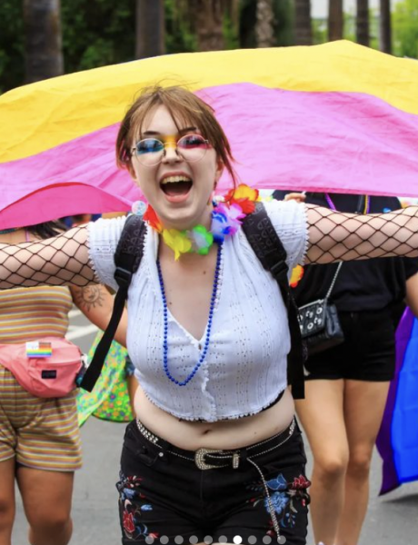 person dressed in colorful clothing with her arms outstretched holding a pansexual flag behind her