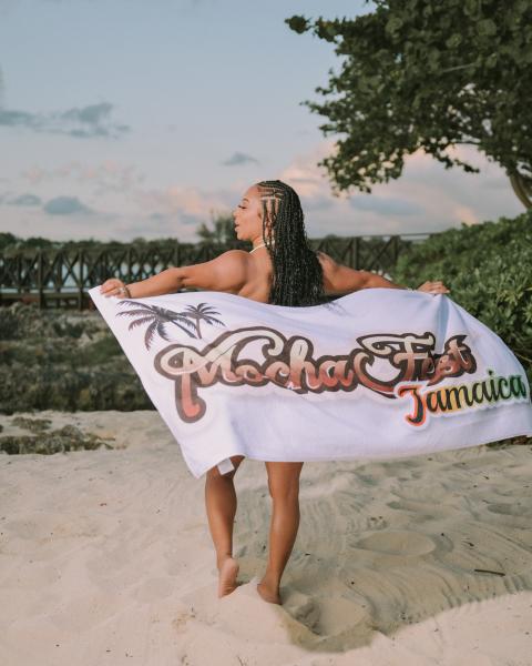 Woman standing on a beach holding a towel with the words 'Mocha Fest Jamaica' behind her