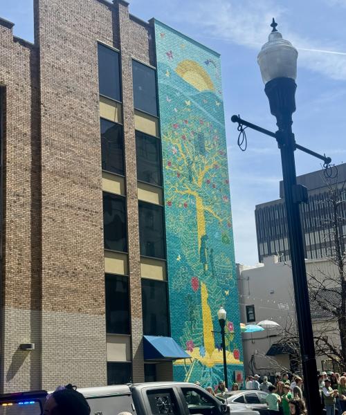 A colorful downtown Huntsville mural featuring a bright yellow tree, flowers, butterflies, and a rising sun stretches up the side of a brick building. People gather along the sidewalk below during a community event, with streetlights and downtown buildings in the background.