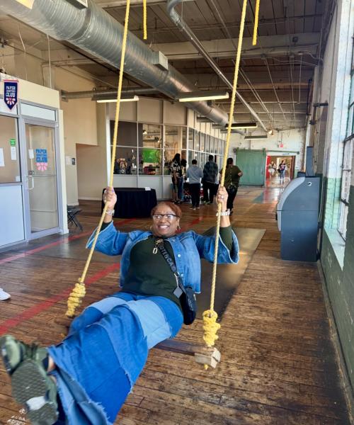 A visitor smiles while swinging on a large rope swing inside Lowe Mill ARTS & Entertainment. The historic mill's wide hallway features exposed ductwork, wood floors, artist studios, and visitors exploring the creative space in the background.