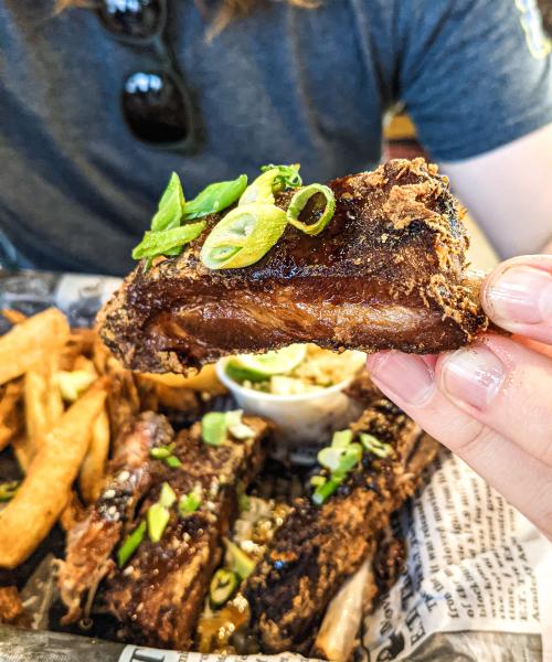 Hand holding a glazed rib topped with green onions, with more ribs and fries in the background at Straight to Ale.