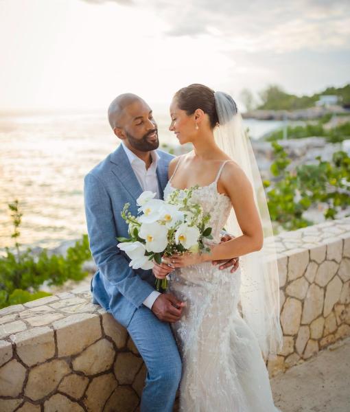 A groom in a blue suit leans against a low wall overlooking the Caribbean Sea at sunset while embracing his bride who is smiling at him.
