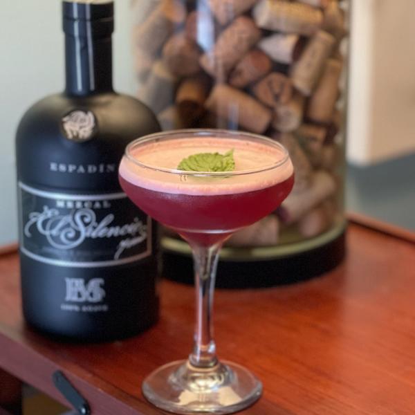 A purple drink in a coup glass sits on a wooden bar.