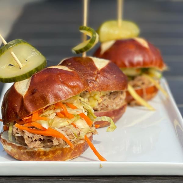 Three small buns topped with pulled pork and cole slaw sit on a white plate. Each has a pickle on a toothpick in the top.