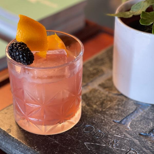 A light pink cocktail in a short glass sits with a blackberry and an orange twist for garnish.