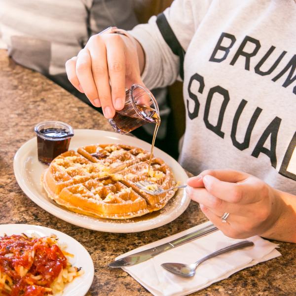 Person wearing a Brunch Squad shirt pouring maple syrup on a Belgian waffle at Cozy Table in Bloomington