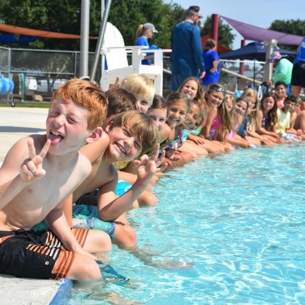 Kids sit on the edge of a swimming pool and smile.