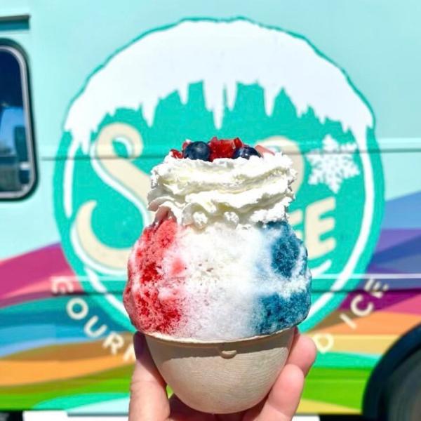 Person holds up snow cone with red, which and blue flavoring and toppings