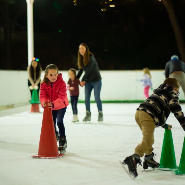 a girl with a cone for support while ice skating and other people ice skating in the background
