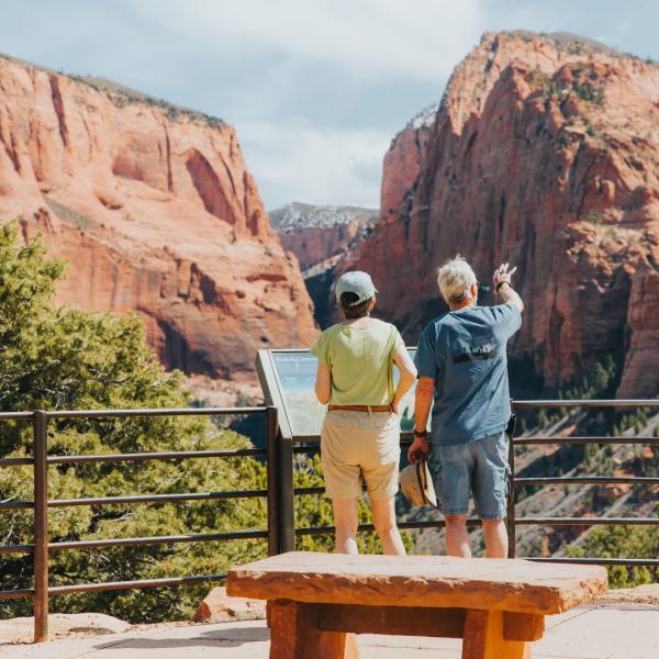 A couple stands at an interpretive sign in Kolob Canyons (north of Zion National Park), pointing at the vibrant orange cliff formations across from the scenic overlook.