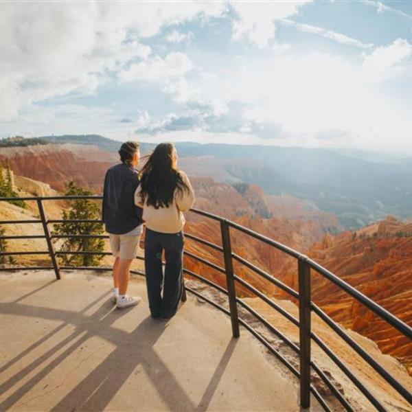A couple stands at a wrought iron fence of the Sunset Overlook in Cedar Breaks National Monument looking out at blue skies and rust orange rock formations in a large natural amphitheater.