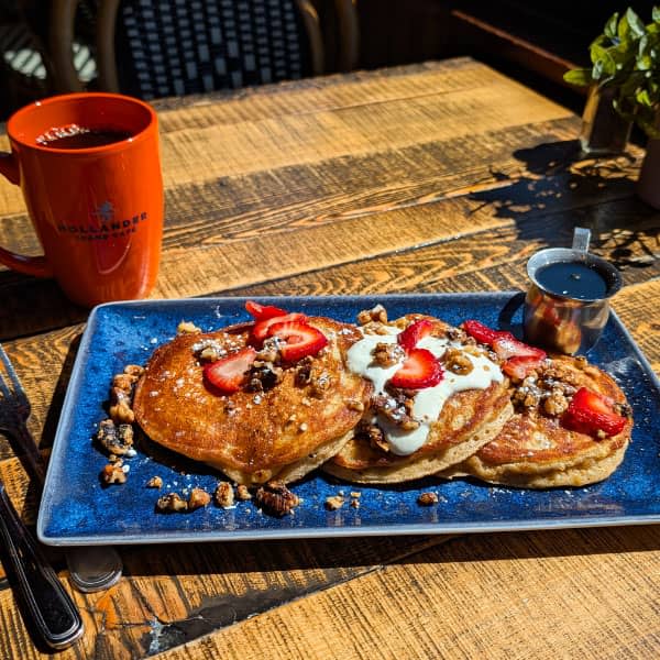 Three pancakes topped with fruit and cream, and a cup of coffee with Cafe Hollander on it.