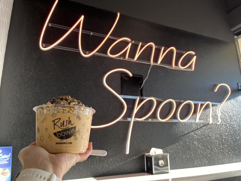 A picture of an acai bowl from Rush Bowls in front of a sign that says "Wanna Spoon?"