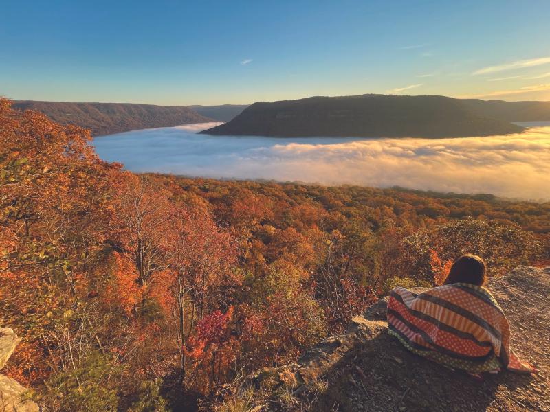 woman sits at Snooper's Rock wrapped in quilt watching sunrise at overlook