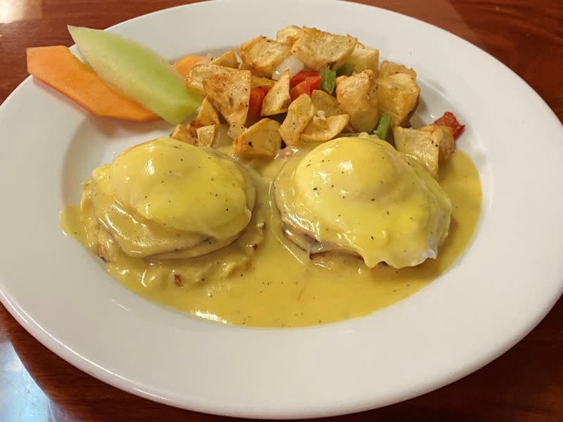 a white plate with fruit, potatoes, and English muffins with eggs and Canadian bacon with a yellow sauce on them