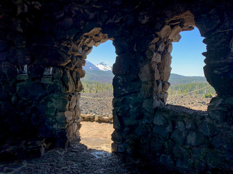 Photo is taken from inside a lava rock fort, looking out through a doorway and window showing snowy mountains in the distance.