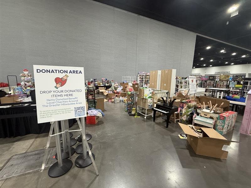 Donation collection area at the end of a trade show with tables, shelves and boxes filled with toys, games, household items and children's products. A sign invites exhibitors to drop off donated items for local charities in the Greater Milwaukee area.Image 3:
