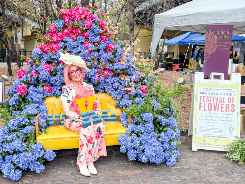 A woman in a floral dress and wide-brim hat sits on a bright yellow bench surrounded by a large arch of blue hydrangeas and pink flowers, holding colorful “MKE” letters. A sign nearby reads “Milwaukee Public Market Festival of Flowers,” with tents and market stalls in the background.