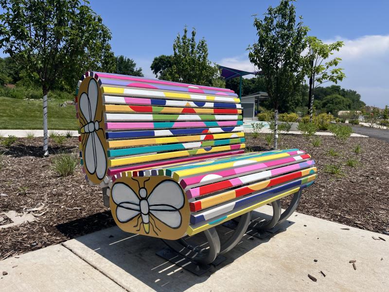 Thin wood panels make up the bench, colored with yellow, indigo, teal, green, and pink. On the sides of the bench, two large animated fireflies are featured.