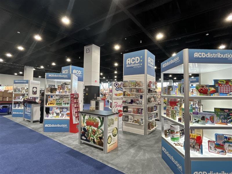 Trade show floor inside a large convention hall with carpeted aisles and multiple toy and gift product displays. ACD Distribution booths feature shelves stocked with games, collectibles and retail merchandise under bright overhead lighting.
