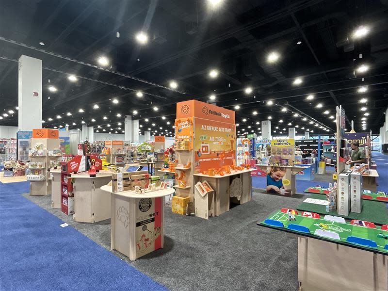 Exhibit booth featuring educational toys, children's products and games displayed on shelves and demonstration tables. The booth sits within a busy trade show floor surrounded by other exhibitors.