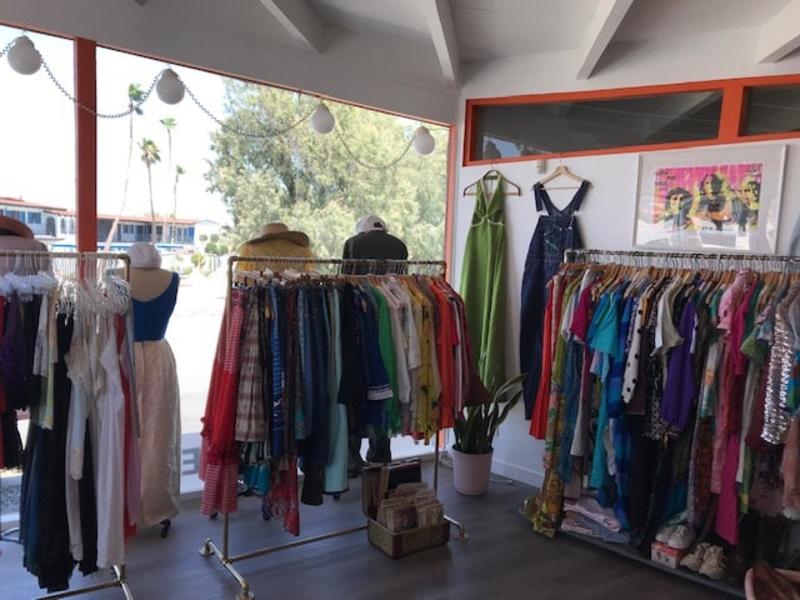 A boutique interior with colorful clothing on racks, a few mannequin displays with dresses and hats, a plant, and a view outside showing palm trees.