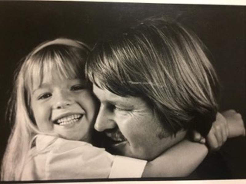 Black and whtie photo of a young girl with blonde hair and bangs smiling at the camera with her arms wrapped around her father's neck. He is looking off from the camera and smiling.