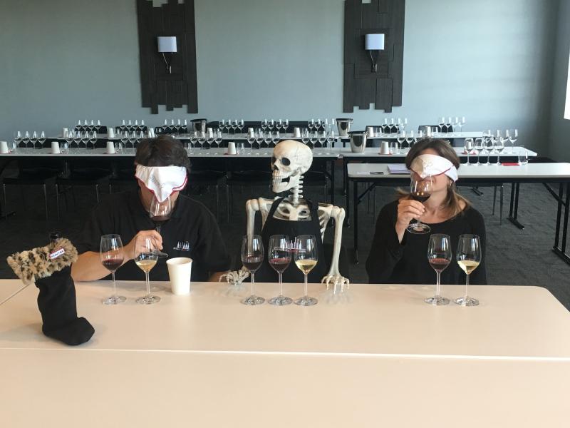 Two people blindfolded tasting a glass of wine with a fake skeleton, "Bob" sitting in between them.