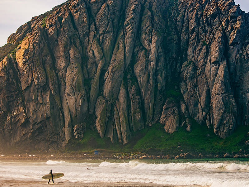 A surfer holding his board walks on the shore near Morro Rock