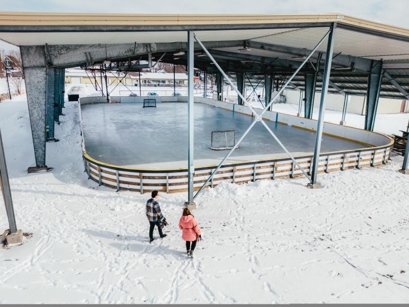 Fisherville Lions Park’s outdoor covered ice rink surrounded by snow on a bright winter day. Two people, one wearing a plaid jacket and the other in a pink coat, walk toward the rink carrying their skates. The rink has hockey nets set up and is partially sheltered by a metal roof with open sides, revealing nearby houses and parked cars in the background.